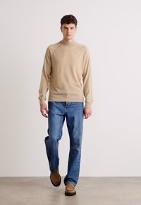Beige høyhalset genser med ribbestrikkede mansjetter og nederkant, sammen med blå jeans og brune sko, vist mot en nøytral bakgrunn.