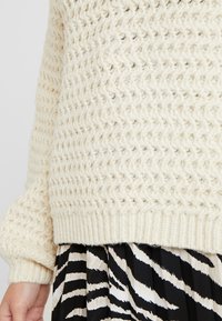 Cremfarbener gestrickter Pullover mit einem strukturierten Zickzackmuster, gerippten Bündchen und Saum. getragen über einem plissierten Rock mit schwarz-weißen Zebramustern.