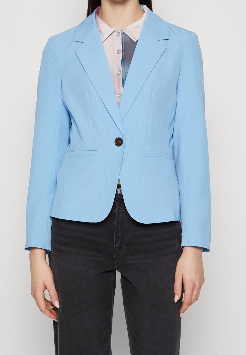 Femme portant un blazer bleu clair à un seul bouton sur une chemise rose pâle à col et un pantalon noir, debout devant un fond uni.