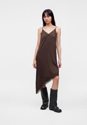 Jeune femme debout portant une robe marron asymétrique sans manches avec un bord en dentelle et des bottes noires montant jusqu'aux genoux sur un fond uni.