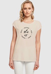 Beige kortærmet t-shirt med cirkulært sort tekstdesign. Sætningerne inkluderer "hun er", "mor" og "mama", med en afslappet pasform og blød tekstur.