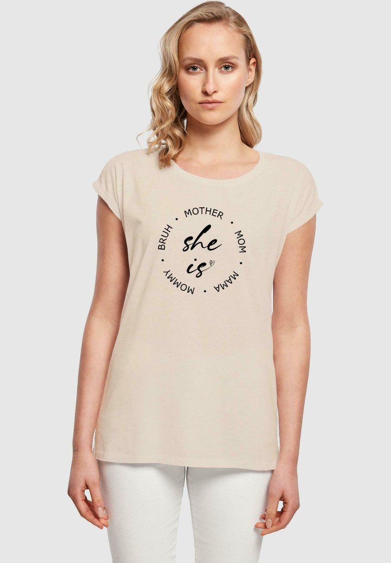 Beige kortærmet t-shirt med cirkulært sort tekstdesign. Sætningerne inkluderer "hun er", "mor" og "mama", med en afslappet pasform og blød tekstur.