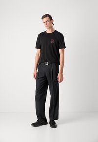 Zwart katoenen t-shirt met rood "HUGO" logo, gecombineerd met zwarte maatwerk broek en zwarte schoenen, staand tegen een neutrale achtergrond.