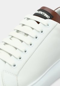 Λευκό δερμάτινο αθλητικό παπούτσι με καφέ και μαύρες λεπτομέρειες, διαθέτει επίπεδα λευκά κορδόνια, κυκλικές οπές και υφασμένο πάτο. Λογότυπο της μάρκας στη γλώσσα.