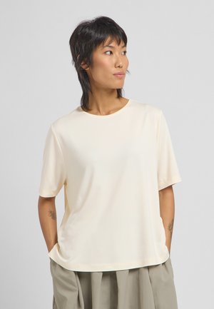 Lichtbeige t-shirt met korte mouwen en een ronde halslijn, met een relaxte pasvorm, gemaakt van soepel stof, gedragen over losse grijze broeken.