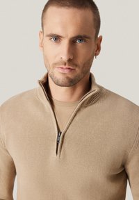 Beżowy sweter dziany z kołnierzem na półzamek i prążkowanymi detalami. Tkanina wydaje się miękka, o subtelnej fakturze, z minimalistycznym wzornictwem.