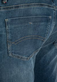 Vaqueros de mezclilla azul oscuro con un solo bolsillo trasero que presenta un detalle de costura curva. La textura parece suave y ligeramente desgastada.