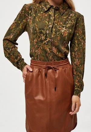Femme portant un chemisier vert à manches longues à fleurs, rentré dans une jupe en cuir marron taille haute avec taille à cordon.