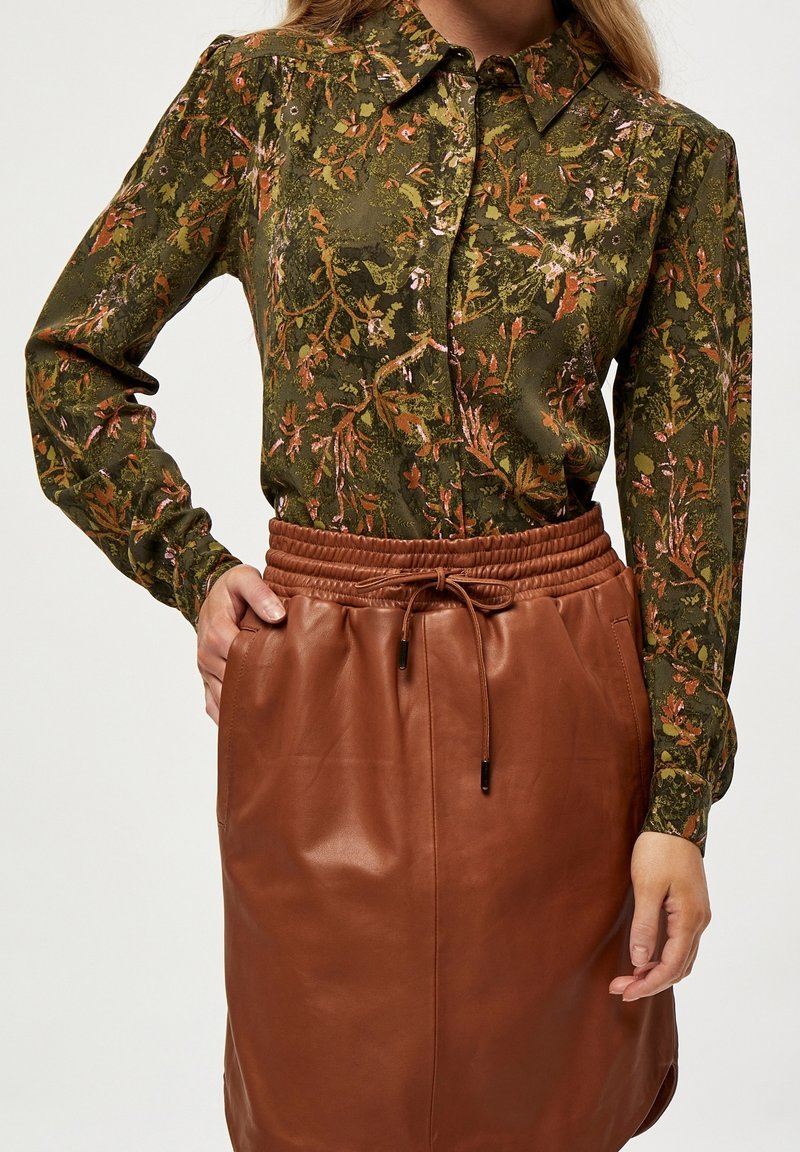 Femme portant un chemisier vert à manches longues à fleurs, rentré dans une jupe en cuir marron taille haute avec taille à cordon.