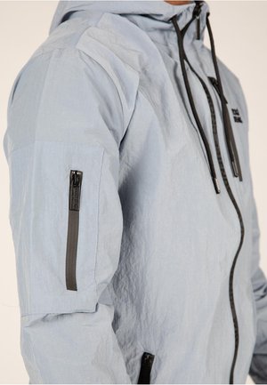 Vaaleansininen huppari, jossa on tekstitetty kangas, mustat vetoketjut ja yksityiskohdat, vinosti sijoitettu vetoketjupussi vasemmassa hihassa.