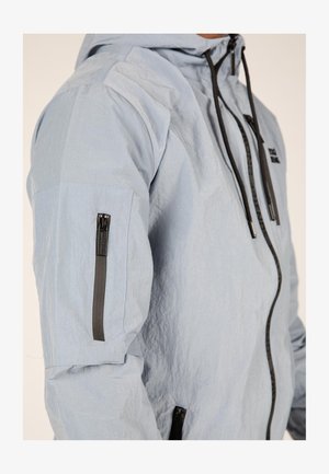 Vaaleansininen huppari, jossa on tekstitetty kangas, mustat vetoketjut ja yksityiskohdat, vinosti sijoitettu vetoketjupussi vasemmassa hihassa.