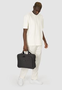 Bolsa para portátil de tela gris con asas dobles, cierre de cremallera y bolsillo frontal. El modelo lleva puesta una camiseta blanca y pantalones claros.