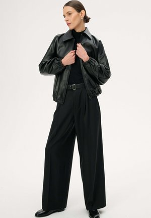 Kobieta z zaczesanymi do tyłu włosami, nosząca czarną skórzaną kurtkę, szerokie czarne spodnie i czarne buty, poprawiająca suwak kurtki.