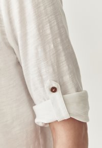 Langärmeliges Hemd aus leicht beigem Stoff, mit umschlagbaren Manschetten und einem weißen Band sowie einem braunen Knopfdetail zur Befestigung.
