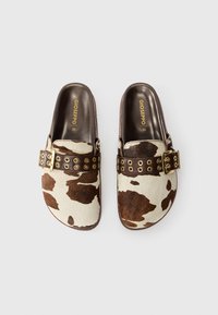 Kuhfell-Mules mit braun- und cremefarbenem gepunktetem Muster, ausgestattet mit einem breiten braunen Riemen mit goldenen Ösen und Schnallendetails. Größe 36.