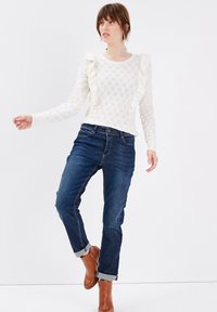 Pull en tricot blanc avec des accents volants sur les côtés, associé à un jean bleu foncé retroussé aux chevilles et des bottines brunes. Fond lumineux.