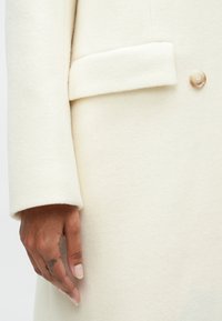 Manteau en laine couleur crème avec une couture d'épaule, des poignets repliés et une fermeture à bouton unique à l'avant. Texture lisse et design minimal.