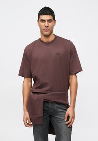 Mladý muž nosí hnedé oversized tričko s opásanou mikinou okolo pása a tmavošedé džínsy, stojí proti jednému pozadiu.