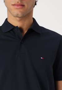 Tumedat sinise värvi polo särk, mis on valmistatud puuvillast, kahe nööbiga krae, lühikesed varrukad ja rinnal väike punase, valge ning sinise värvi logo.