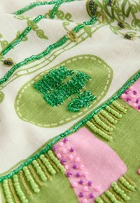 Detailní záběr na látku se zelenými, růžovými a fialovými korálky přišitými na zelený a bílý textil s vyšitými vzory.
