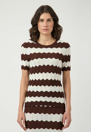 Femme aux cheveux foncés mi-longs, vêtue d'un haut marron et blanc à manches courtes avec des rayures ondulées et d'une jupe assortie, debout sur un fond neutre.