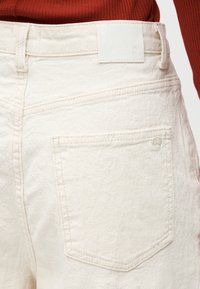 Κοντινή λήψη τσέπης πίσω και λεπτομέρεια ζώνης σε κρεμ χρωματιστό παντελόνι με υφή, φορεμένο με σκουρόχρωμο κόκκινο ριγωτό τοπ.