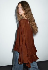 Blusa marrone a maniche lunghe con design a ruffle a strati. Tessuto leggero e fluido. Abbinata a jeans blu chiaro su uno sfondo neutro.