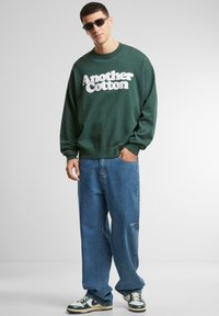 Sudadera verde con texto esponjoso blanco que dice "Otro Algodón", combinada con jeans azules holgados y gafas de sol negras. Zapatillas con detalles verdes.