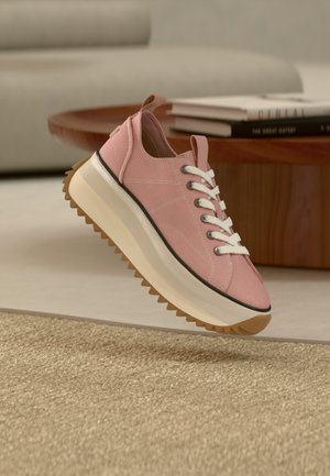 Baskets à plateforme roses avec lacets blancs et semelle épaisse beige à nervures, posées au bord d'un tapis beige, avec une table en bois et des livres en arrière-plan.