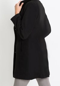 Svart kappa i mjukt tyg, med figurnära passform, knäppning fram och sidofickor. Långa ärmar och slät textur.