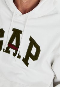 Bílá mikina s velkými olivově zelenými písmeny "GAP" a červeným nápisem "Athletic", s kapucí na stahovací šňůrku a měkkou texturou.