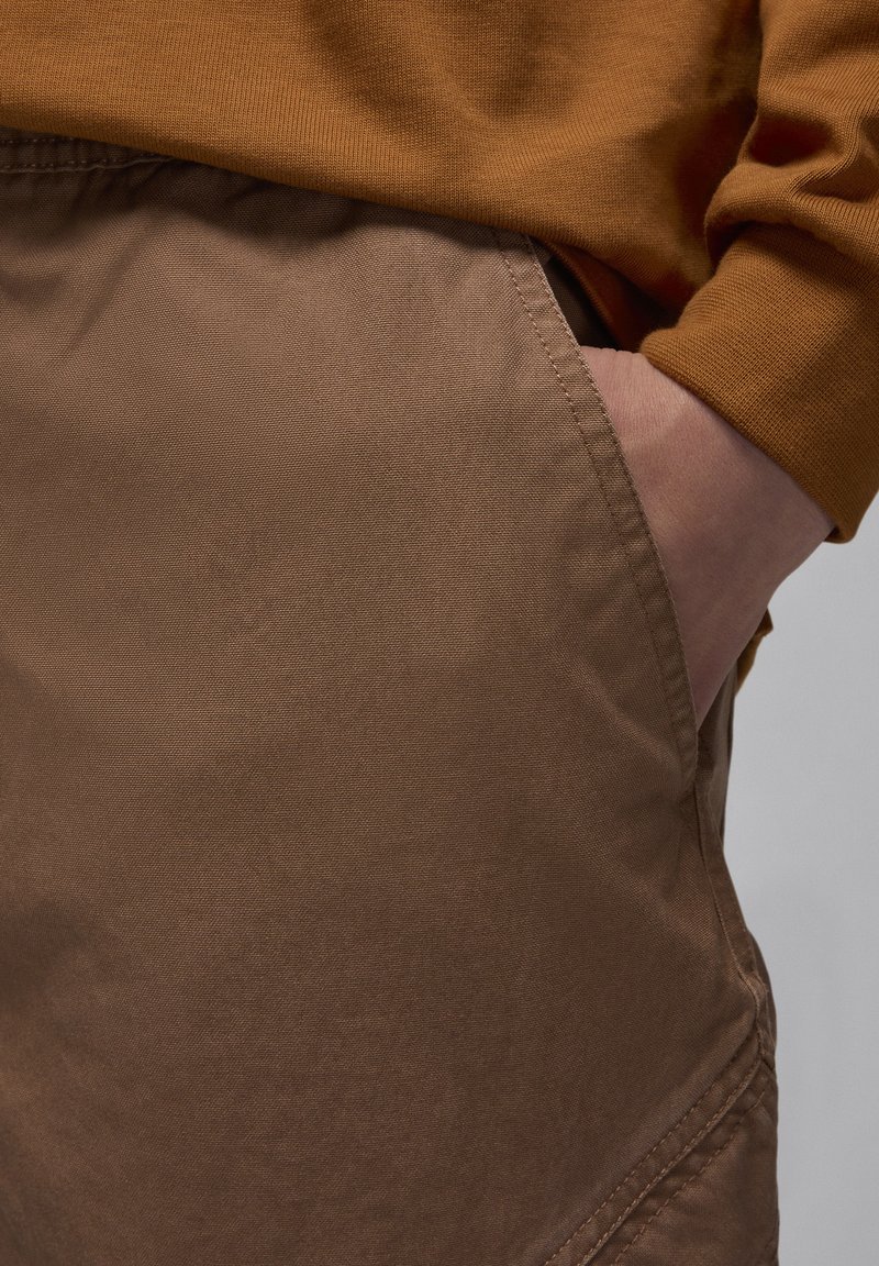 Pantaloni in tessuto leggero marrone con tasche laterali visibili e una texture sottile. La parte superiore mostra il polsino di una camicia a maniche lunghe marrone.