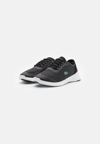 Une paire de chaussures de sport noires à lacets avec des semelles blanches et un petit logo de crocodile vert sur le côté, présentée sur un fond blanc uni.