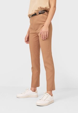 Frau trägt schlanke Kamelhautfarbene Hose mit schwarzem Gürtel und weißen Turnschuhen, steht vor einfachem hellem Hintergrund.