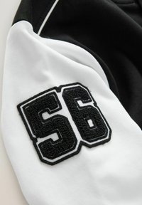 Indumento in bianco e nero con un grande numero ricamato "56" sulla manica, caratterizzato da sezioni di tessuto lisce e testurizzate in contrasto.