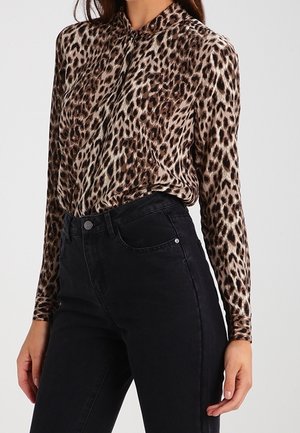 Kobieta nosząca bluzkę z długim rękawem w panterkę, wsuniętą w czarne spodnie z wysokim stanem na jednolitym tle.