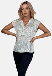 Biała bluzka w czarne plamki, dekolt w kształcie V, rękawy na ramionach i luźny krój. Materiał wydaje się gładki z delikatnym połyskiem.