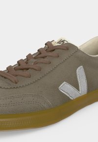 Baskets décontractées beiges en suédine avec lacets marron, logo "V" blanc cousu sur le côté, et semelle extérieure en caoutchouc marron translucide.