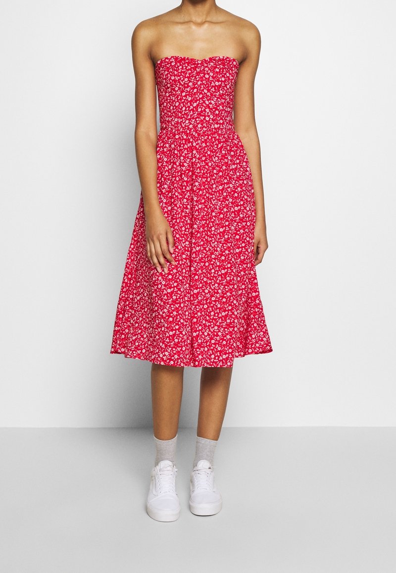 Robe rouge sans bretelles à motif floral, corsage ajusté et jupe évasée, associée à des baskets blanches et des chaussettes grises.