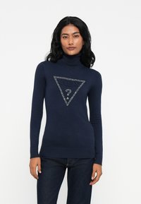 Mujer con cabello negro largo que lleva un su�éter de cuello alto azul marino con un logo de Guess adornado y jeans oscuros, frente a un fondo blanco.
