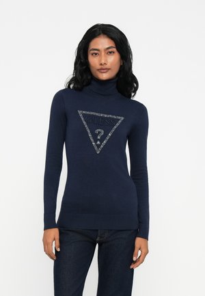 Donna con lunghi capelli neri che indossa un maglione a collo alto blu navy con un logo Guess impreziosito e jeans scuri su uno sfondo bianco.