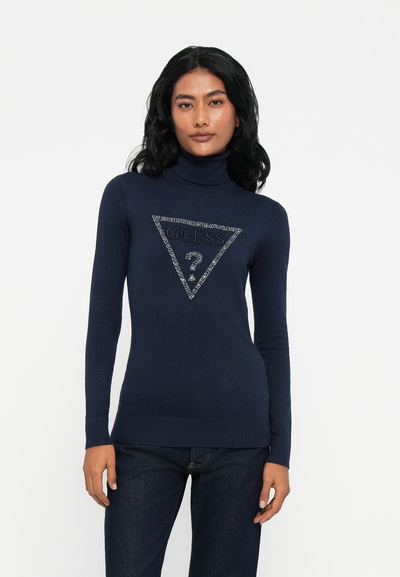 Mujer con cabello negro largo que lleva un suéter de cuello alto azul marino con un logo de Guess adornado y jeans oscuros, frente a un fondo blanco.