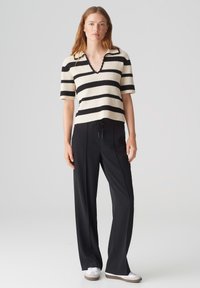 Gestreiftes Strick-Polohemd in Creme und Schwarz, kurze Ärmel, V-Ausschnitt mit welligem Rand, kombiniert mit schwarzen Jogginghosen und weißen Schuhen.