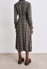 Longue robe à carreaux en olive et noir avec un dos à boutons, taille ceinturée et manches longues, associée à des bottines brunes.
