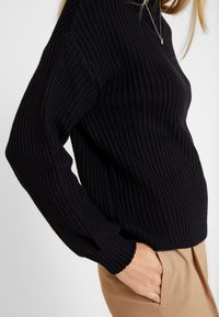 Black knitted sweater with long sleeves and textured fabric; ribbed cuffs and hem; paired with light beige trousers.