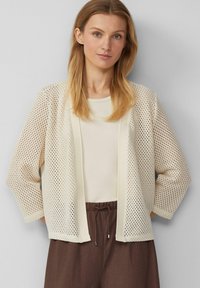 Leichter, cremefarbener Cardigan mit Netzdesign, offenem Vorderteil und dreiviertellangen Ärmeln, kombiniert mit einem beigen Oberteil und braunen Hosen.