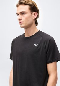 Νέος άνδρας με ανοιχτό καστανό μαλλί, φοράει μαύρο μπλουζάκι Puma, κοιτάζει αριστερά σε απλό ανοιχτό γκρι φόντο.