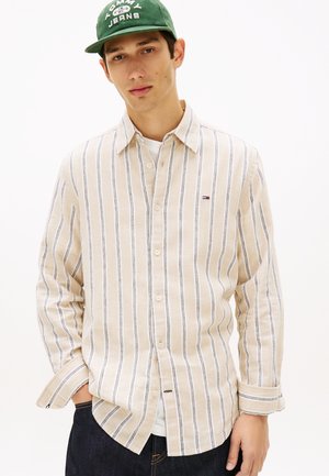 Ung mann iført en beige og hvit skjorte med vertikale striper og knapper, mørke jeans og en grønn Tommy Jeans-caps, stående foran en ensfarget bakgrunn.