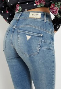 Detailní záběr na osobu v modrých džínách Guess s trojúhelníkovým logem na zadní kapse a květinovým černým topem s bílými puntíky.