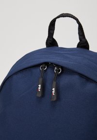Zaino blu navy con una superficie in tessuto liscio, dotato di maniglia superiore e doppie zip con dettaglio del logo.